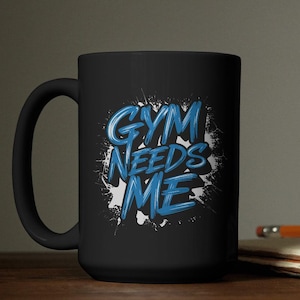 May include: Black ceramic mug with the text "GYM NEEDS ME" in blue, stylized lettering. The text is set against a white splatter design. The mug has a curved handle and is suitable for hot or cold beverages.