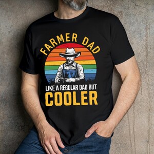 Pu&ograve; includere: T-shirt nera con la scritta "FARMER DAD" sopra un'illustrazione di un contadino con cappello e tuta, sullo sfondo di un tramonto arcobaleno. Il testo "LIKE A REGULAR DAD BUT COOLER" &egrave; sotto.