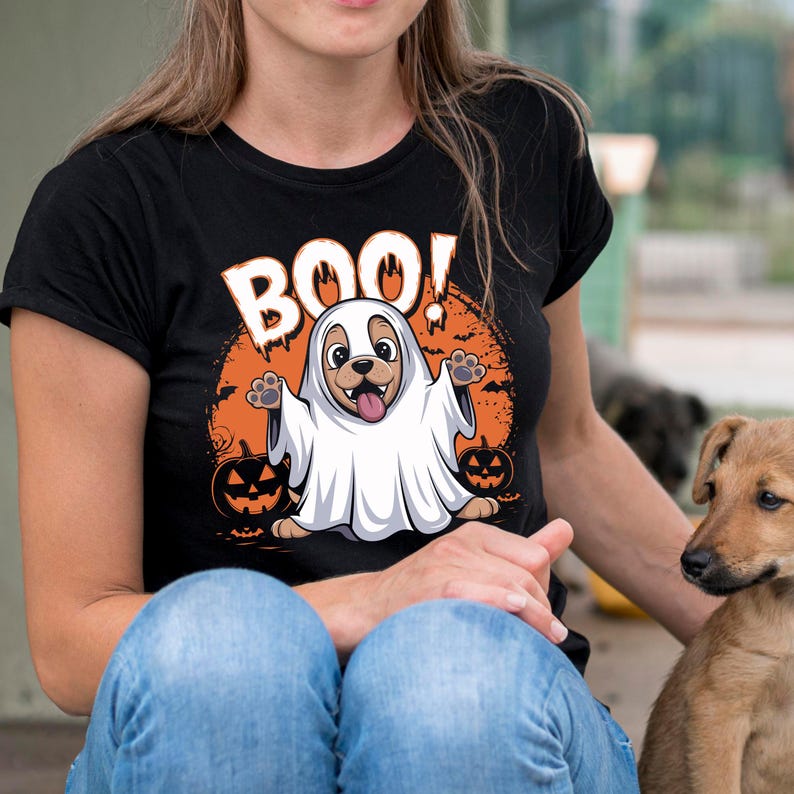 May include: Black t-shirt with a Halloween-themed graphic. The design features a cartoon dog dressed as a ghost with the word "BOO!" above it. Two jack-o'-lanterns are on either side of the dog. The dog is sticking its tongue out.