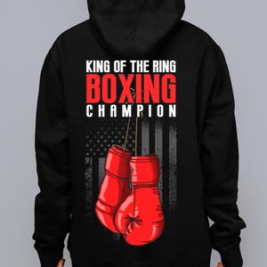 Peut inclure: Sweat-shirt &agrave; capuche noir avec le texte "King of the Ring Boxing Champion" et un graphique de gants de boxe rouges devant un drapeau am&eacute;ricain.