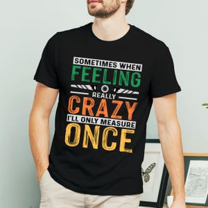 May include: Black t-shirt with a humorous graphic. The text reads "Sometimes when feeling crazy, I'll only measure once." The text is in green, orange, and yellow. The shirt is a crew neck.