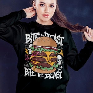 Pu&ograve; includere: Felpa nera con una grafica di un grande hamburger con il testo "Bite the Beast" in lettere bianche e nere.