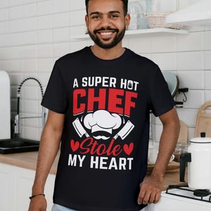 May include: Black t-shirt with the text "A SUPER HOT CHEF Stole MY HEART" in red and white. The design includes a chef's hat, mustache, and crossed cleavers. The shirt is worn by a person in a kitchen setting.