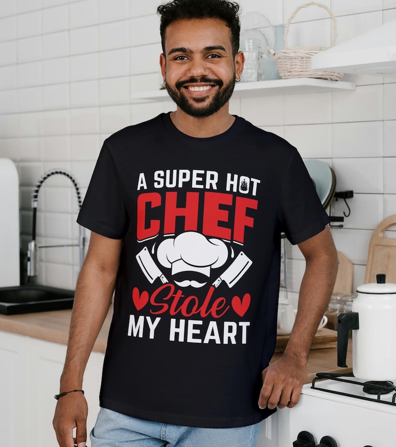 May include: Black t-shirt with the text "A SUPER HOT CHEF Stole MY HEART" in red and white. The design includes a chef's hat, mustache, and crossed cleavers. The shirt is worn by a person in a kitchen setting.