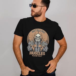 May include: Black t-shirt featuring a graphic of a hooded skeleton holding dumbbells, with the text "INSTALLING MUSCLES PLEASE WAIT" in a circular design. The design is in shades of beige, gray, and white.
