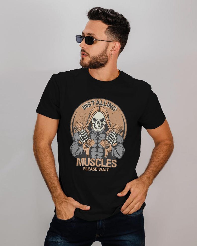 May include: Black t-shirt featuring a graphic of a hooded skeleton holding dumbbells, with the text "INSTALLING MUSCLES PLEASE WAIT" in a circular design. The design is in shades of beige, gray, and white.