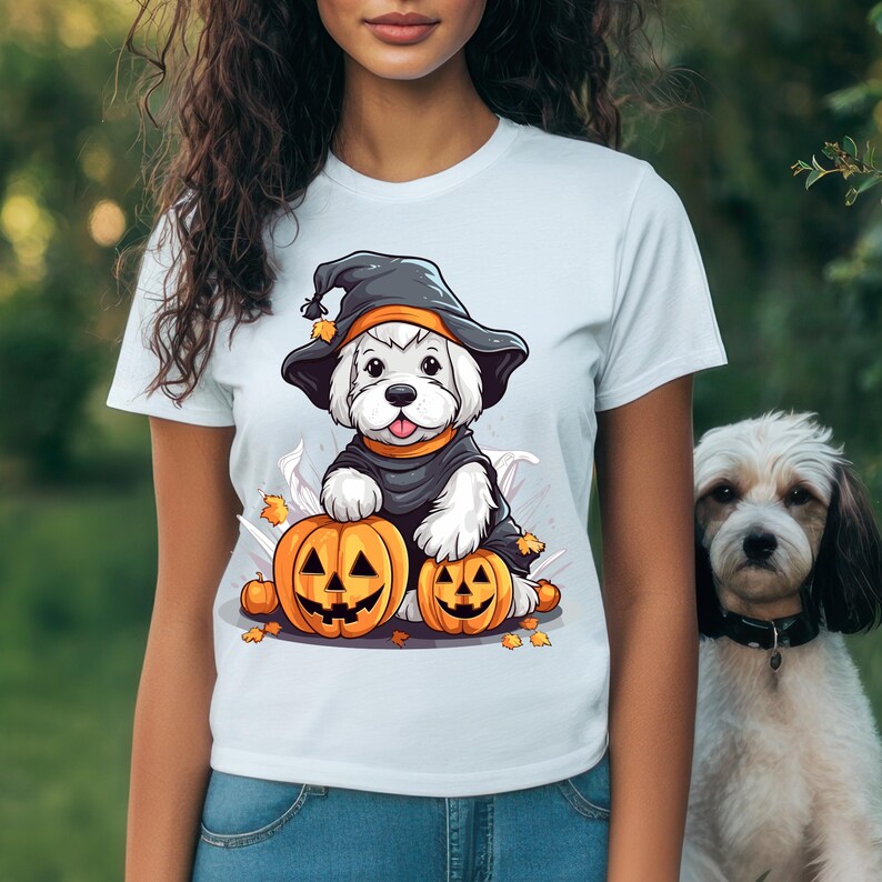 May include: White t-shirt featuring a cartoon dog dressed in a witch costume, sitting on two carved pumpkins. The dog wears a black hat with orange trim and a black cloak. A second dog is visible in the background.
