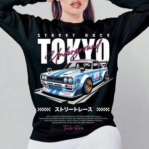Puede incluir: Camiseta negra de manga larga con un dise&ntilde;o de coche de carreras azul y blanco, el texto "TOKYO Underground" y "Turbo Race". El dise&ntilde;o incluye texto japon&eacute;s y el n&uacute;mero 23. El dise&ntilde;o de la camiseta est&aacute; inspirado en las carreras callejeras.