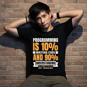 Puede incluir: Camiseta negra con el texto "PROGRAMMING IS 10% WRITING CODE AND 90% UNDERSTANDING WAY IT'S NOT WORKING" en blanco y naranja. La camiseta la lleva una persona.