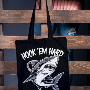 May include: Black tote bag featuring a white shark graphic and the text "HOOK 'EM HARD".