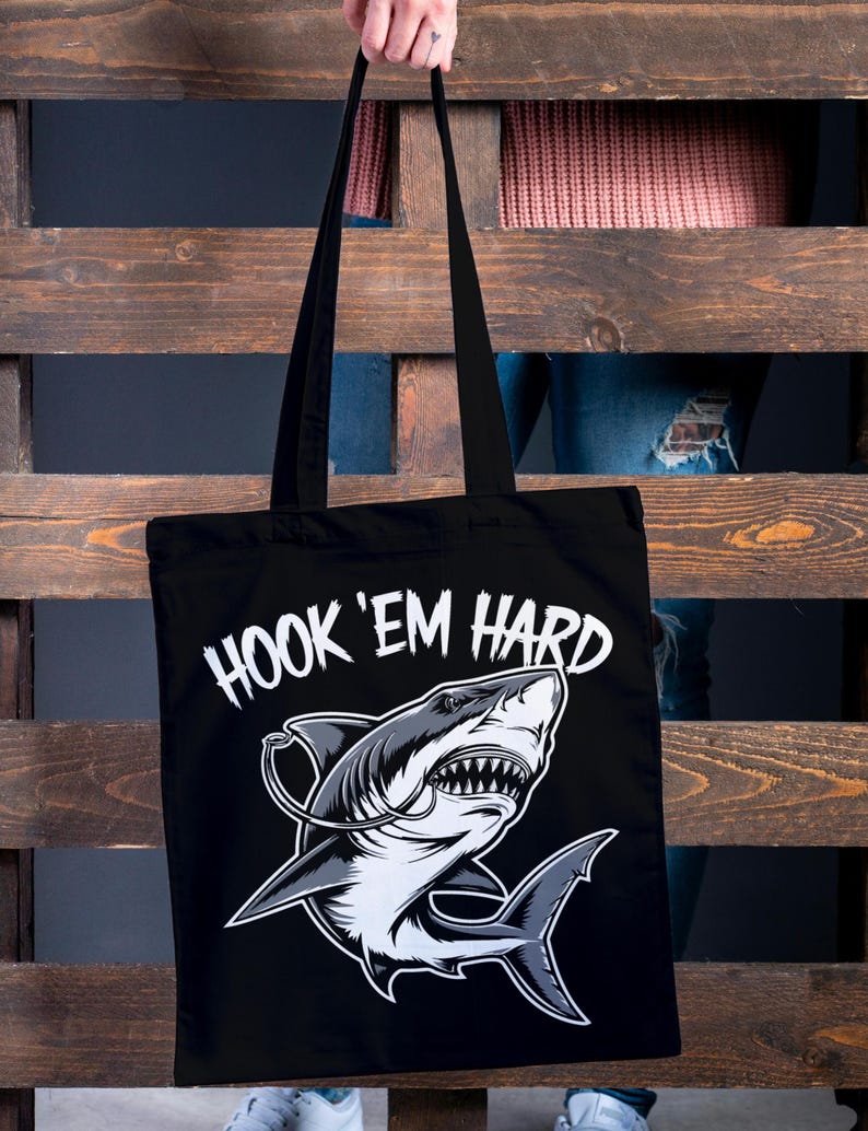 May include: Black tote bag featuring a white shark graphic and the text "HOOK 'EM HARD".