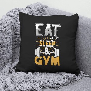 May include: Black square pillow with the words "EAT SLEEP & GYM" in white, gray, and yellow. The design includes a barbell graphic. The pillow is on a gray blanket.