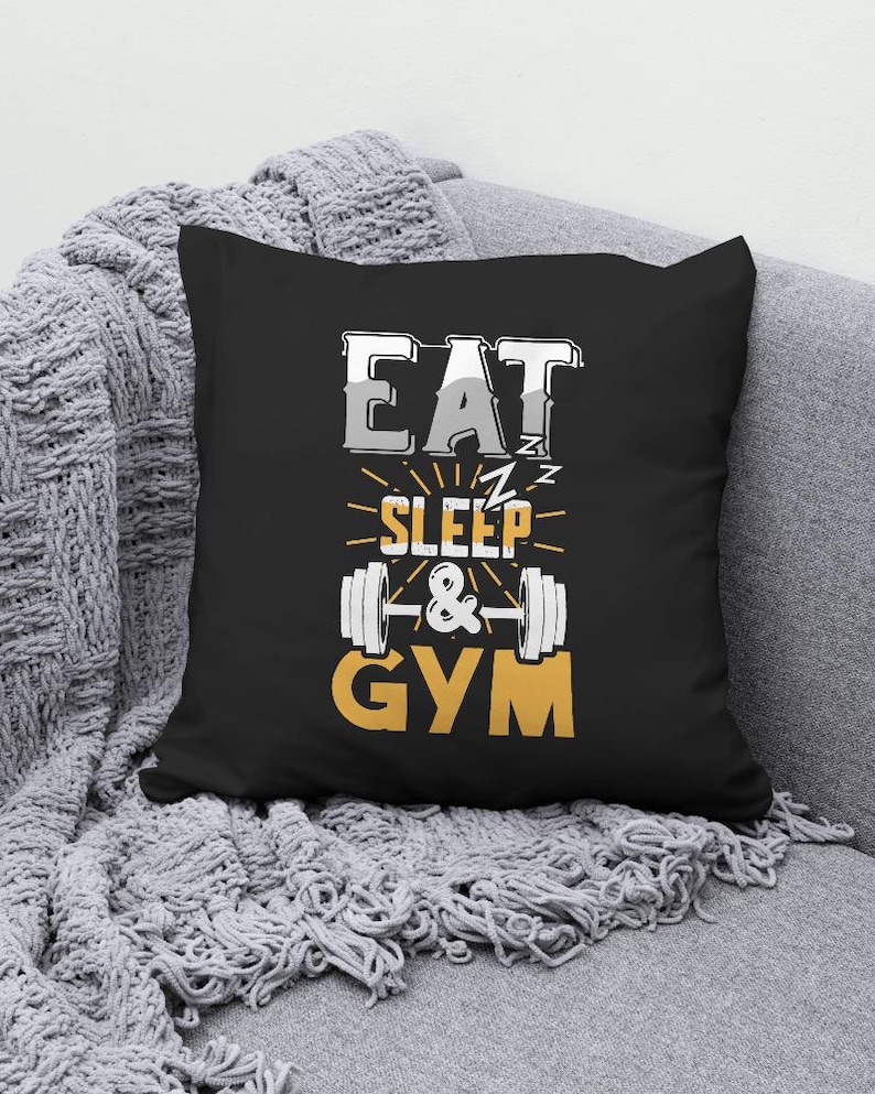 May include: Black square pillow with the words "EAT SLEEP & GYM" in white, gray, and yellow. The design includes a barbell graphic. The pillow is on a gray blanket.