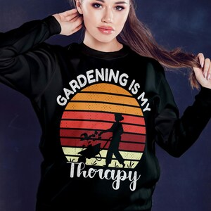 Pu&ograve; includere: Felpa nera con un design grafico raffigurante un tramonto, un giardiniere e la scritta "Gardening is my Therapy" in bianco. Il design include una persona che spinge una carriola e si prende cura delle piante.