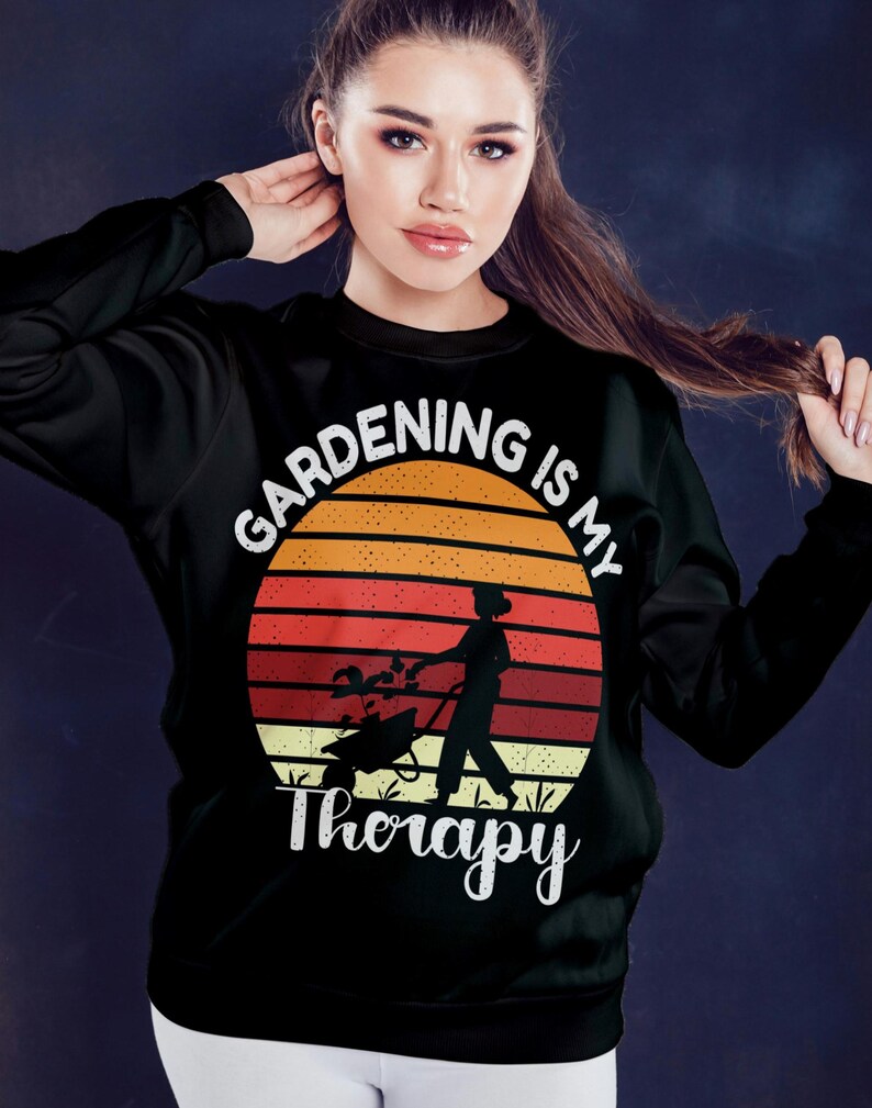 Pu&ograve; includere: Felpa nera con un design grafico raffigurante un tramonto, un giardiniere e la scritta "Gardening is my Therapy" in bianco. Il design include una persona che spinge una carriola e si prende cura delle piante.