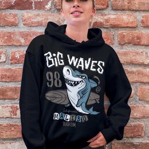 May include: Black hooded sweatshirt featuring a graphic of a shark riding a surfboard. The graphic includes the text "Big Waves 98 California Malibu Beach Team".