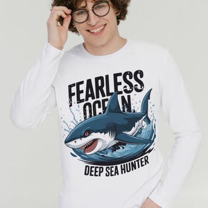 May include: White long-sleeved shirt featuring a graphic of a shark leaping from the water. The text "Fearless Ocean" and "Deep Sea Hunter" is printed on the shirt.