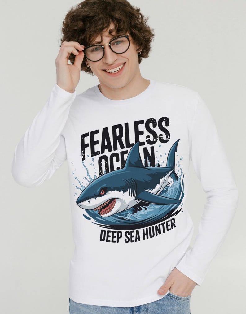 May include: White long-sleeved shirt featuring a graphic of a shark leaping from the water. The text "Fearless Ocean" and "Deep Sea Hunter" is printed on the shirt.