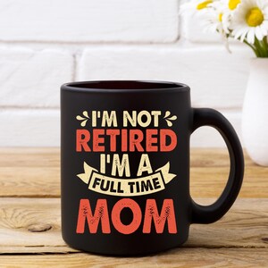 Może przedstawiać: Czarny ceramiczny kubek do kawy z napisem "I'M NOT RETIRED I'M A FULL TIME MOM" w pomarańczowych i białych literach. Kubek ma duży uchwyt i stoi na drewnianej powierzchni.