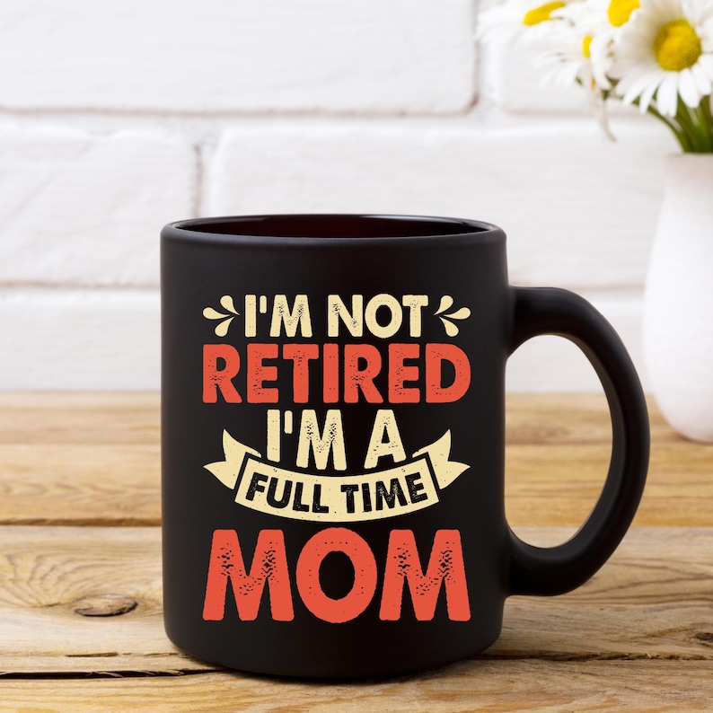 Może przedstawiać: Czarny ceramiczny kubek do kawy z napisem "I'M NOT RETIRED I'M A FULL TIME MOM" w pomarańczowych i białych literach. Kubek ma duży uchwyt i stoi na drewnianej powierzchni.