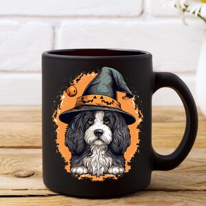 May include: Black ceramic mug featuring a Halloween-themed illustration. The design showcases a Cavalier King Charles Spaniel wearing a witch's hat with a jack-o'-lantern. The dog is set against an orange background with a pumpkin.