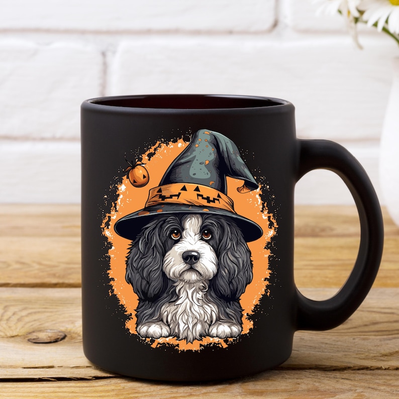 May include: Black ceramic mug featuring a Halloween-themed illustration. The design showcases a Cavalier King Charles Spaniel wearing a witch's hat with a jack-o'-lantern. The dog is set against an orange background with a pumpkin.