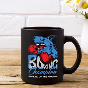 Peut inclure: Une tasse &agrave; caf&eacute; en c&eacute;ramique noire avec un graphique d'un requin bleu portant des gants de boxe. Le texte "Boxing Champion - King of the Ring" est imprim&eacute; sur la tasse.