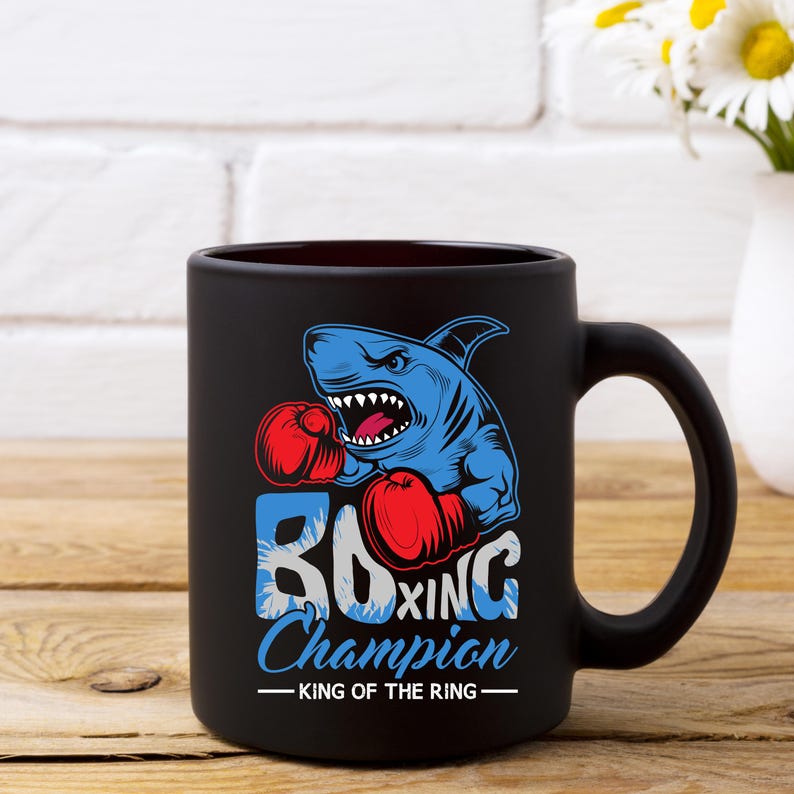 Peut inclure: Une tasse &agrave; caf&eacute; en c&eacute;ramique noire avec un graphique d'un requin bleu portant des gants de boxe. Le texte "Boxing Champion - King of the Ring" est imprim&eacute; sur la tasse.