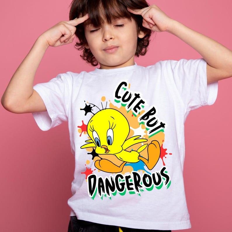 Puede incluir: Camiseta blanca con un dise&ntilde;o de dibujos animados de Tweety Bird y el texto "Cute But Dangerous". El p&aacute;jaro amarillo tiene ojos azules y pies naranjas. El dise&ntilde;o incorpora salpicaduras y estrellas de colores.