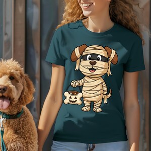 May include: Teal t-shirt featuring a cartoon dog dressed as a mummy, holding a bone-shaped candy bucket. The dog has brown ears and a black eye mask. The bucket has a jack-o'-lantern face. A golden doodle dog is visible on the left.