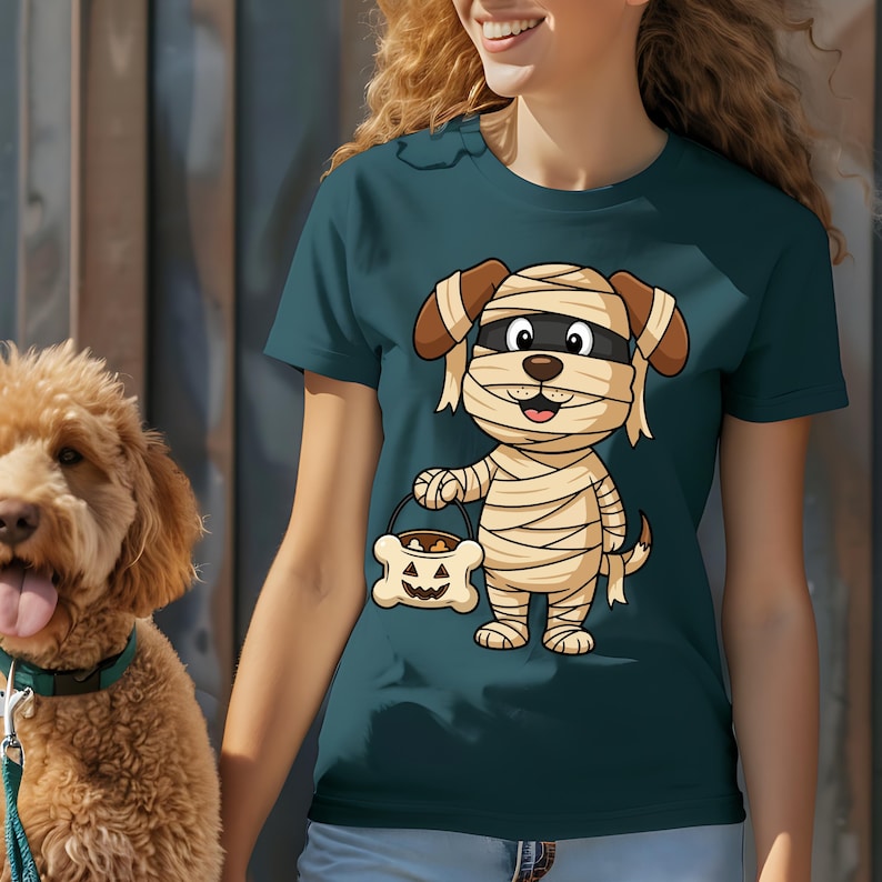 May include: Teal t-shirt featuring a cartoon dog dressed as a mummy, holding a bone-shaped candy bucket. The dog has brown ears and a black eye mask. The bucket has a jack-o'-lantern face. A golden doodle dog is visible on the left.