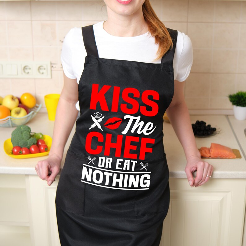 May include: Black apron with the text "KISS The CHEF OR EAT NOTHING" in red and white. The apron has a chef's hat and crossed utensils graphic. The apron is worn by a person in a white shirt.