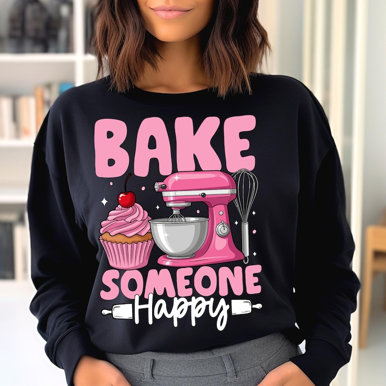 May include: Black sweatshirt featuring a pink graphic with the words "BAKE SOMEONE HAPPY." The design includes a pink stand mixer, a cupcake with a cherry, and a whisk. The word "Happy" is in cursive.