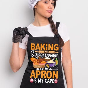 May include: Black apron with the text "BAKING IS MY SUPERPOWER AND MY APRON IS MY CAPE." The apron features illustrations of baking tools and baked goods, including a rolling pin, bread, and cupcakes. The apron is worn by a person wearing a white chef's hat.