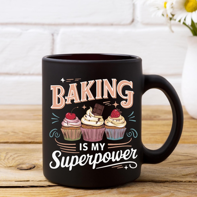 May include: Black ceramic mug with the words "BAKING IS MY SUPERPOWER" in white and tan lettering. The mug features three cupcakes with different toppings. The mug is on a wooden surface.