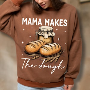 May include: Brown sweatshirt featuring the text "MAMA MAKES" above an illustration of two loaves of bread, a jar of honey, and wheat stalks. The text "The dough" is written below the illustration.