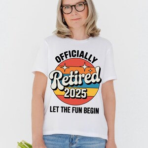Może przedstawiać: Biała koszulka z kolorową grafiką w stylu retro z napisem "OFFICIALLY Retired 2025". Projekt zawiera tło przypominające zach&oacute;d słońca i słowa "LET THE FUN BEGIN" poniżej. Koszulka jest noszona przez osobę.