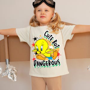 Puede incluir: Una camiseta color crema con un dibujo animado de Tweety Bird y el texto "Cute But Dangerous". El p&aacute;jaro amarillo lleva pantalones cortos azules y est&aacute; rodeado de salpicaduras de pintura coloridas. La camiseta es de manga corta.