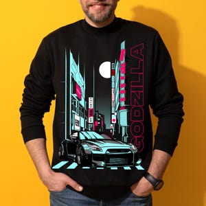 Puede incluir: Sudadera negra con una ilustraci&oacute;n estilizada de un coche deportivo plateado en una ciudad nocturna. El dise&ntilde;o incluye la palabra "GODZILLA" en rosa, una luna llena y edificios con detalles azules y rosas.
