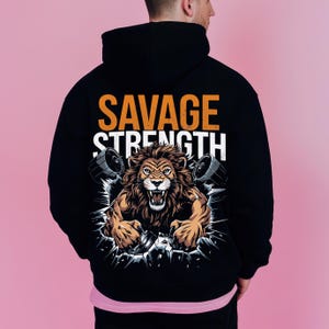 Puede incluir: Sudadera con capucha negra con un estampado gr&aacute;fico de un le&oacute;n rompiendo una pared con el texto "SAVAGE STRENGTH" en naranja.