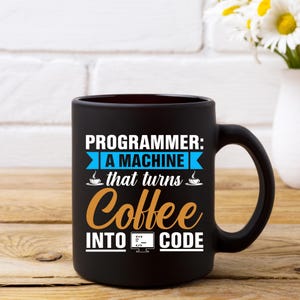 Puede incluir: Taza de caf&eacute; negra con el texto "PROGRAMMER: A MACHINE that turns Coffee INTO CODE" en blanco, azul y naranja. La taza tiene un asa grande y est&aacute; sobre una superficie de madera.