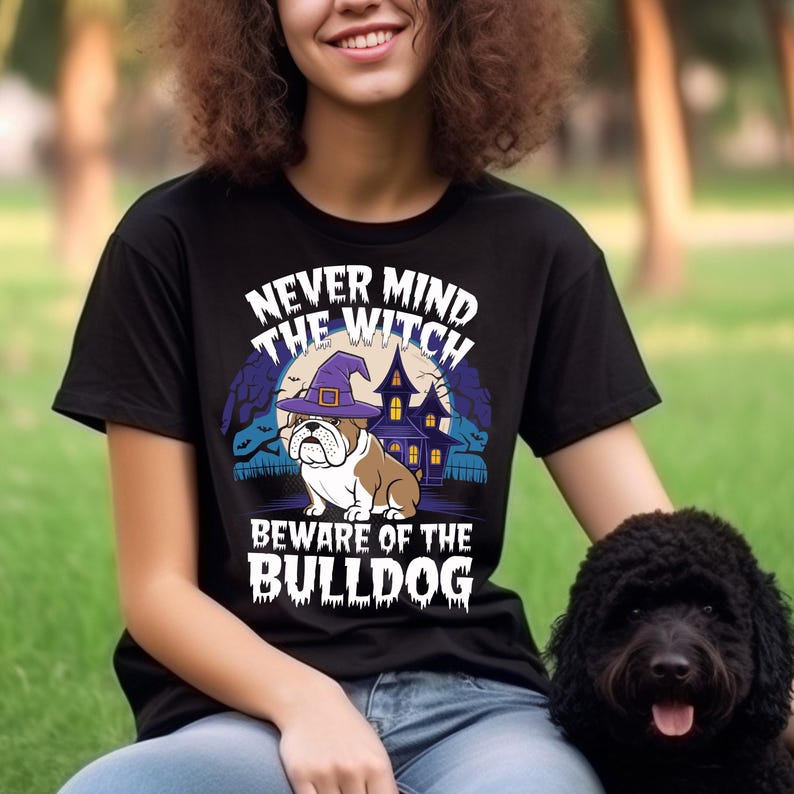 May include: Black t-shirt with a Halloween-themed graphic. The design features a bulldog wearing a witch hat in front of a haunted house, with the text "NEVER MIND THE WITCH BEWARE OF THE BULLDOG". A black dog is in the image.