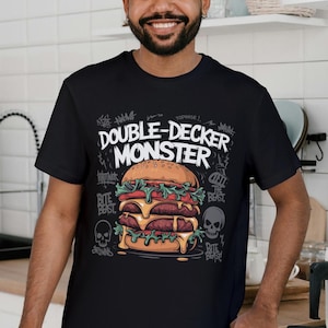 Pu&ograve; includere: Maglietta nera con una grafica di un grande hamburger a due piani con il testo "Double-Decker Monster" in bianco. L'hamburger ha lattuga, pomodoro, formaggio e due hamburger di manzo. La grafica &egrave; circondata da testo in stile graffiti e teschi.