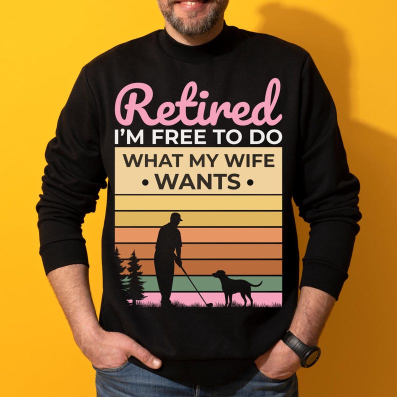 Może przedstawiać: Czarna bluza z napisem "Retired I'M FREE TO DO WHAT MY WIFE WANTS" w kolorze r&oacute;żowym i białym. Grafika przedstawia golfistę z psem i tłem zachodu słońca. Bluza ma okrągły dekolt.
