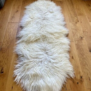 May include: A white sheepskin rug with long, fluffy fur. The rug is laid out on a wooden floor.