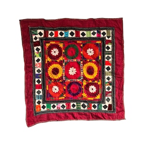 May include: A square, embroidered textile with a deep red border. The central design features nine colourful squares with floral patterns in red, yellow, and white. The border includes black and white diamond shapes and a black trim.