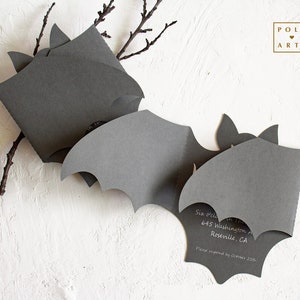 May include: A black bat-shaped invitation with the text "Six o'clock in the evening" and an address. The invitation is folded in half and has a white background.