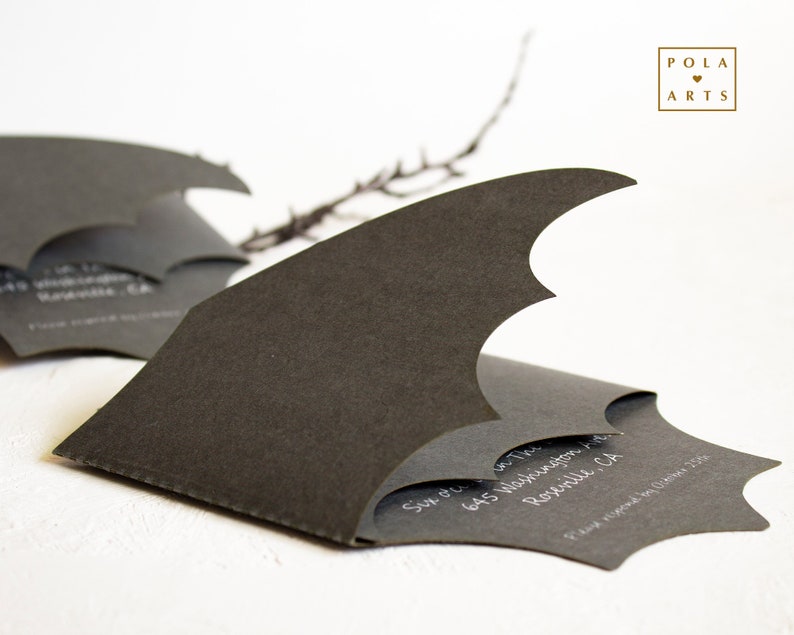 May include: A black bat-shaped invitation card with white text that reads "Sun Oct 1st, 645 Washington Rd,  Roseville, CA.  Please respond by October 1st, 2023."  The card is folded in half and the top flap is folded back to reveal the text.  The card is on a white surface.