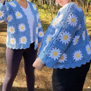 K&ouml;nnte beinhalten: Zwei blaue geh&auml;kelte Strickjacken mit wei&szlig;en G&auml;nsebl&uuml;mchen-Designs. Die Jacken haben einen gewellten Saum und sind aus weichem Garn gefertigt.