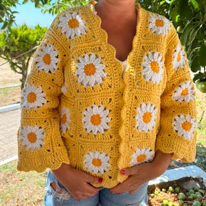 May include: A long-sleeved crochet cardigan made of yellow square panels, each featuring a white daisy with a bright yellow center. The garment has a V-neckline, a scalloped front edge, and a textured, hand-stitched appearance throughout the design.